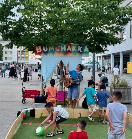 Kinder spielen in der Ballbande auf einem Platz und One Take Toni zeigt auf sein Bumchakka schild