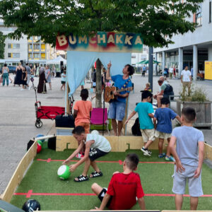 Kinder spielen in der Ballbande auf einem Platz und One Take Toni zeigt auf sein Bumchakka schild