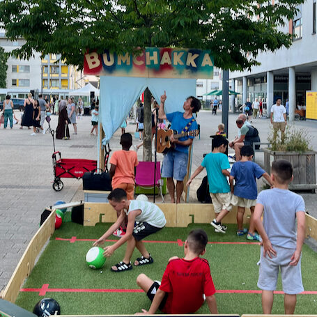 Kinder spielen in der Ballbande auf einem Platz und One Take Toni zeigt auf sein Bumchakka schild