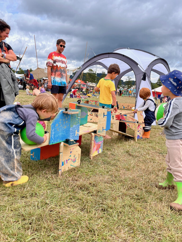 Kinder in Matschhosen auf der Wiese eines Festivalgeländes. Sie Spielen mit bunten Bällen und haben aus kleinen Toren und Teilen der Ballbande eine Ballbahn gebaut