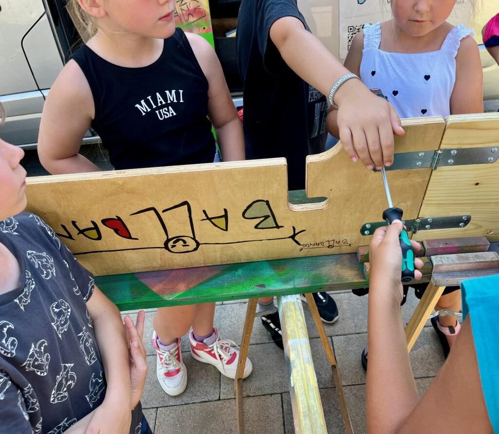 ein Kind hält das Scharnier fest ein anderes schraubt eine Schraube fest