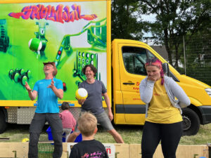 drei Menschen stehen vor einem grossen bunten Bus, einem Spielmobil und werfen bunte Bälle in die Luft, lachen
