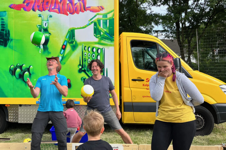 drei Menschen stehen vor einem grossen bunten Bus, einem Spielmobil und werfen bunte Bälle in die Luft, lachen
